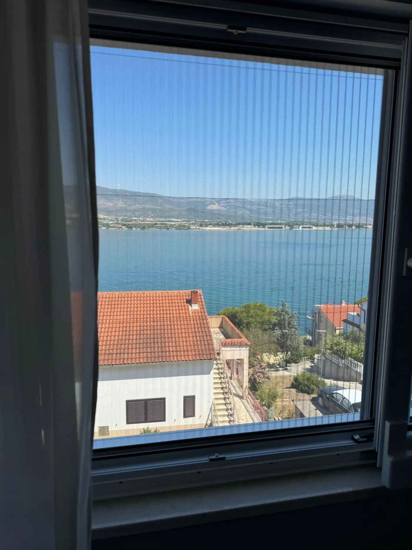 Komarnik u tamnom okviru prozora, kroz mrežu pogled na more i krovove
